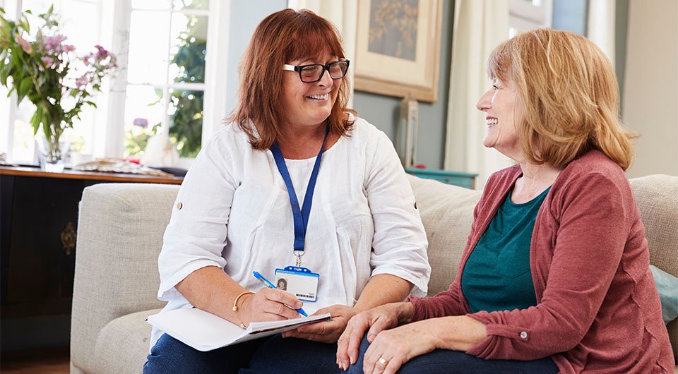 Two women are seated on a couch, engaged in a conversation, with one taking notes on a clipboard.