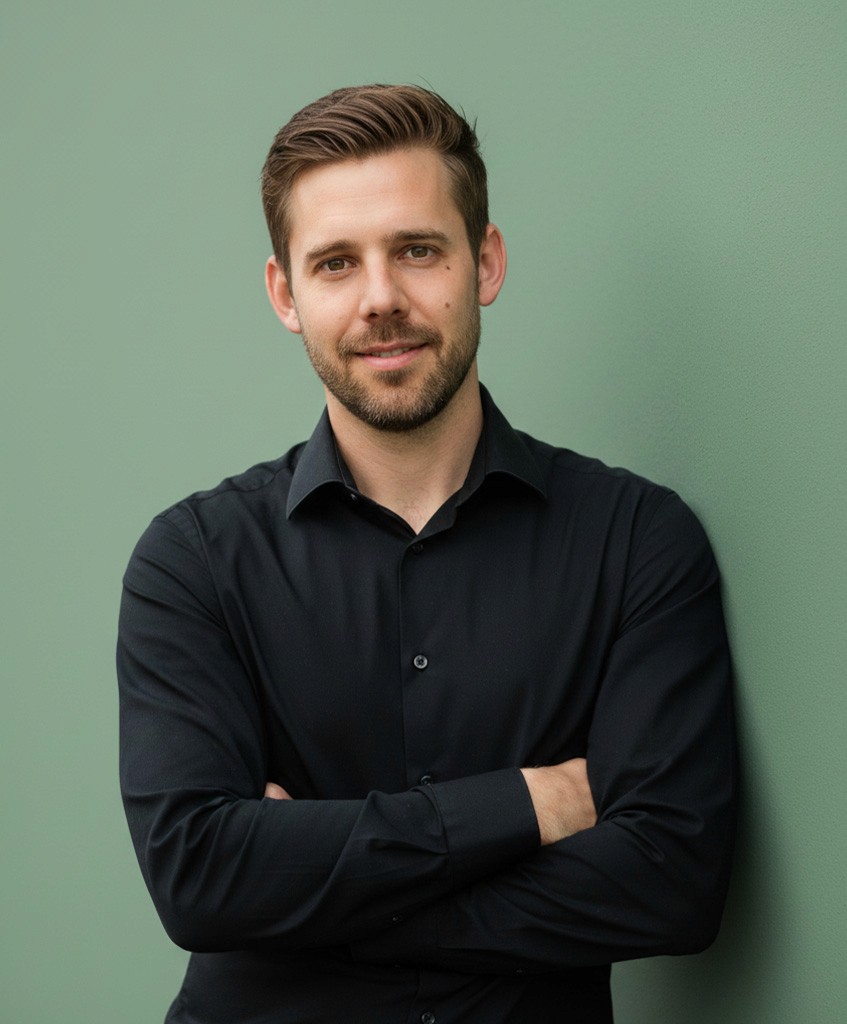 A young person with curly hair smiles at the camera while sitting in front of a laptop against a dark background.