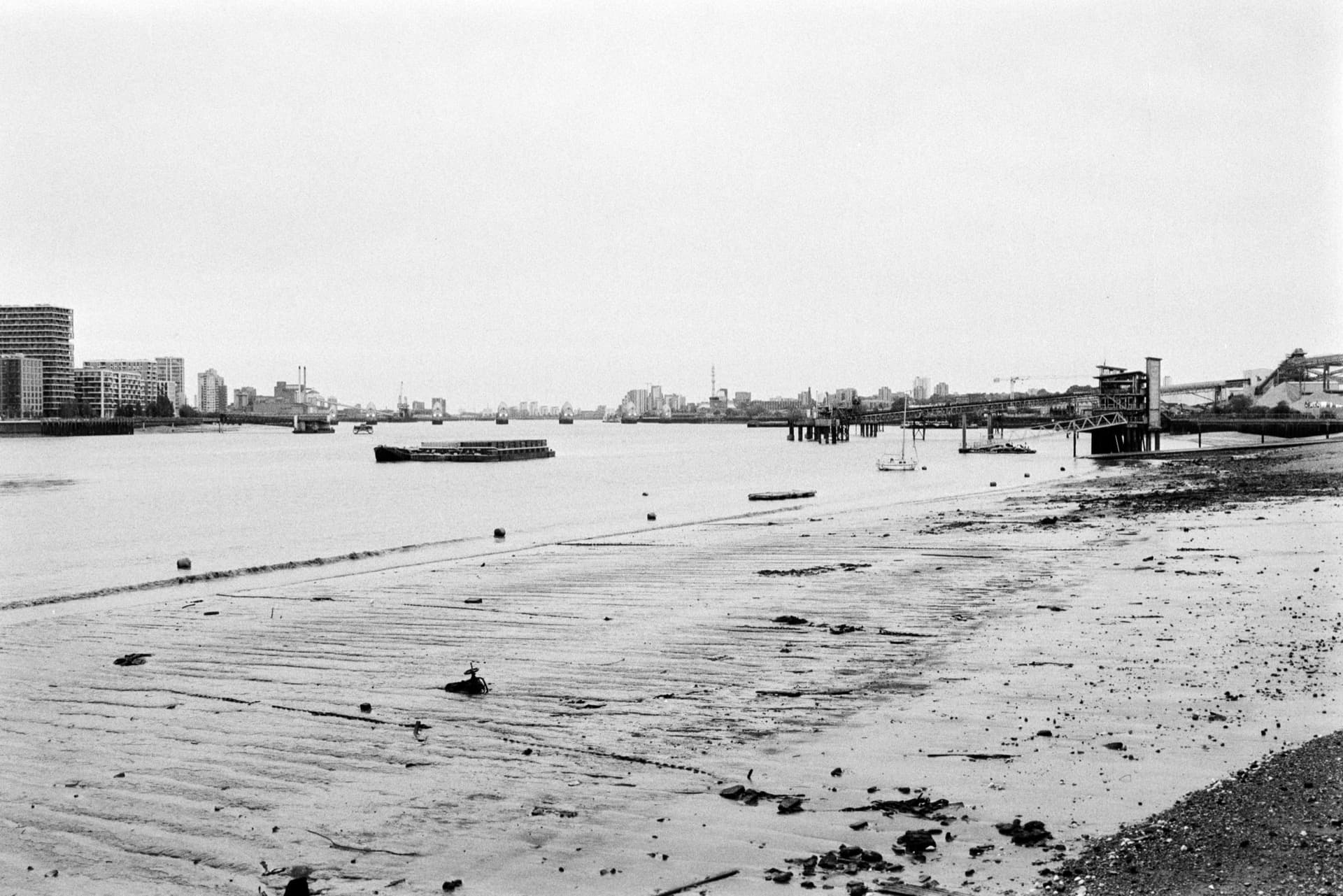 Thames foreshore looking toward Thames Barrier, exposed mudflats at low tide with debris, barges and vessels on water, industrial piers and distant development visible on both banks