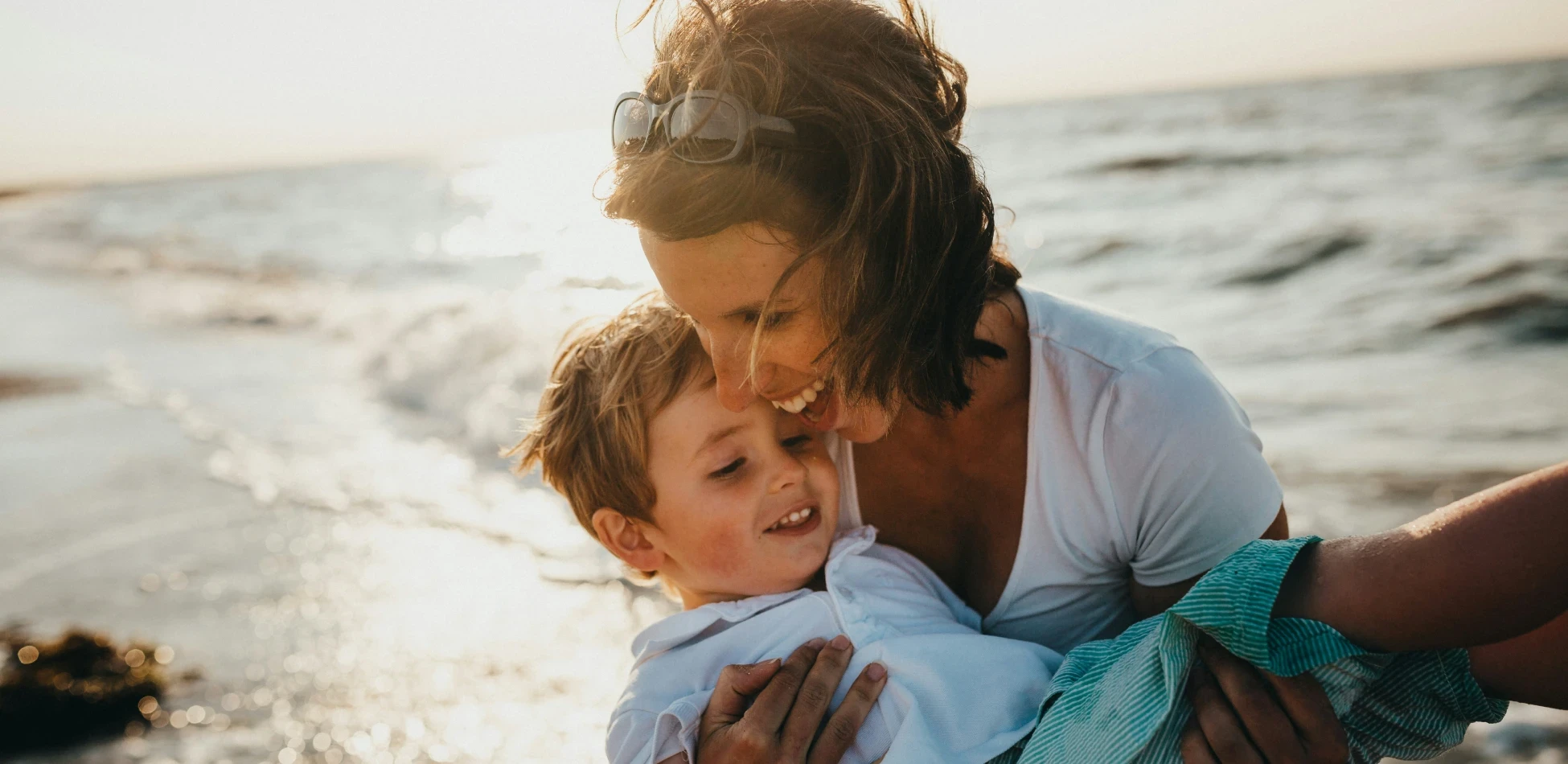 Mother and young son embracing at sunset on beach, captured in warm golden light showing emotional connection.
