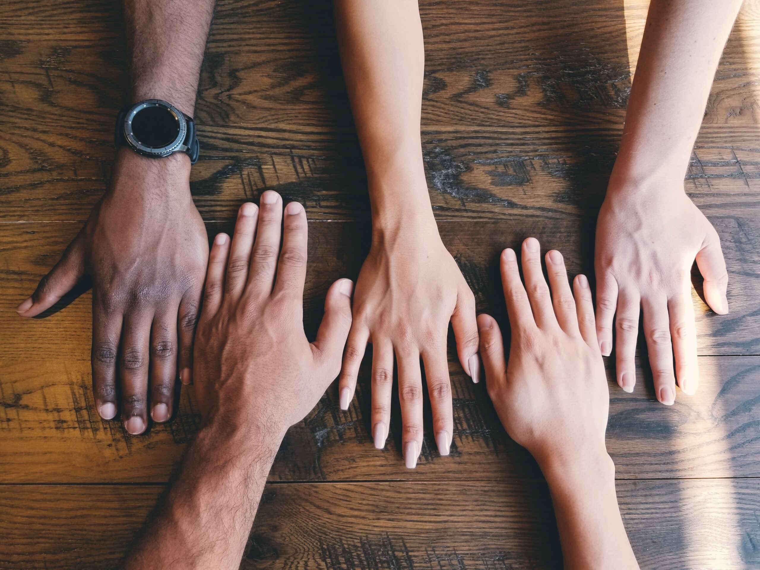 Multiple hands of diverse skin tones resting palm-down on a wooden table
