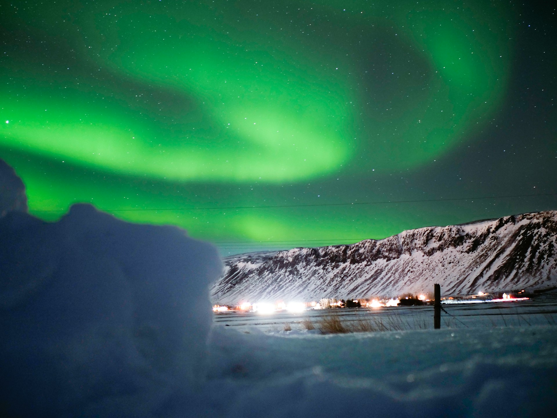 Green northern lights swirling above snow-covered mountains and a winter valley in Iceland on a clear night.