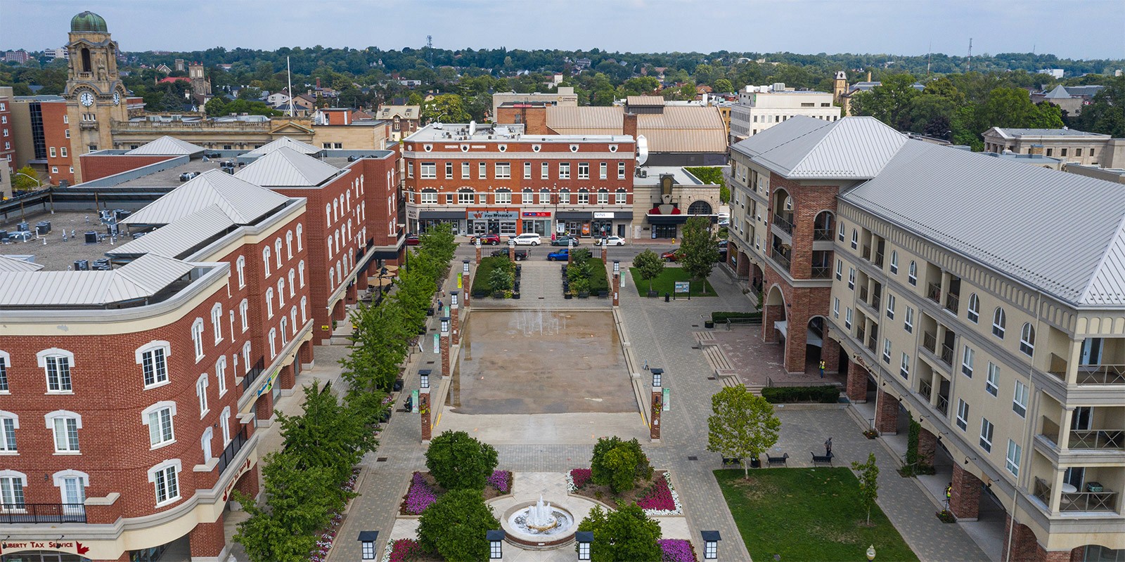 Aerial view of a pedestrian plaza surrounded by multi-story buildings with red brick and beige facades, featuring a central water fountain, symmetrical garden beds with pink flowers, and a shallow reflecting pool with fountains. The area is framed by tree-lined walkways and leads toward a street with shops, cars, and a historic clock tower in the background.