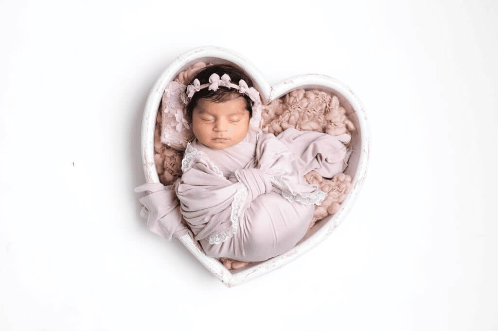 Peacefully sleeping newborn wrapped in soft fabric, posed safely in a heart-shaped bowl on a cosy pink blanket