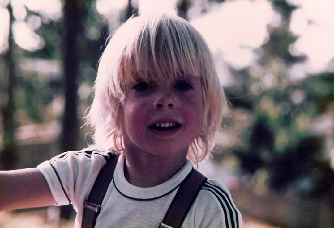 niño con pelo largo rubio en el bosque con tirantes