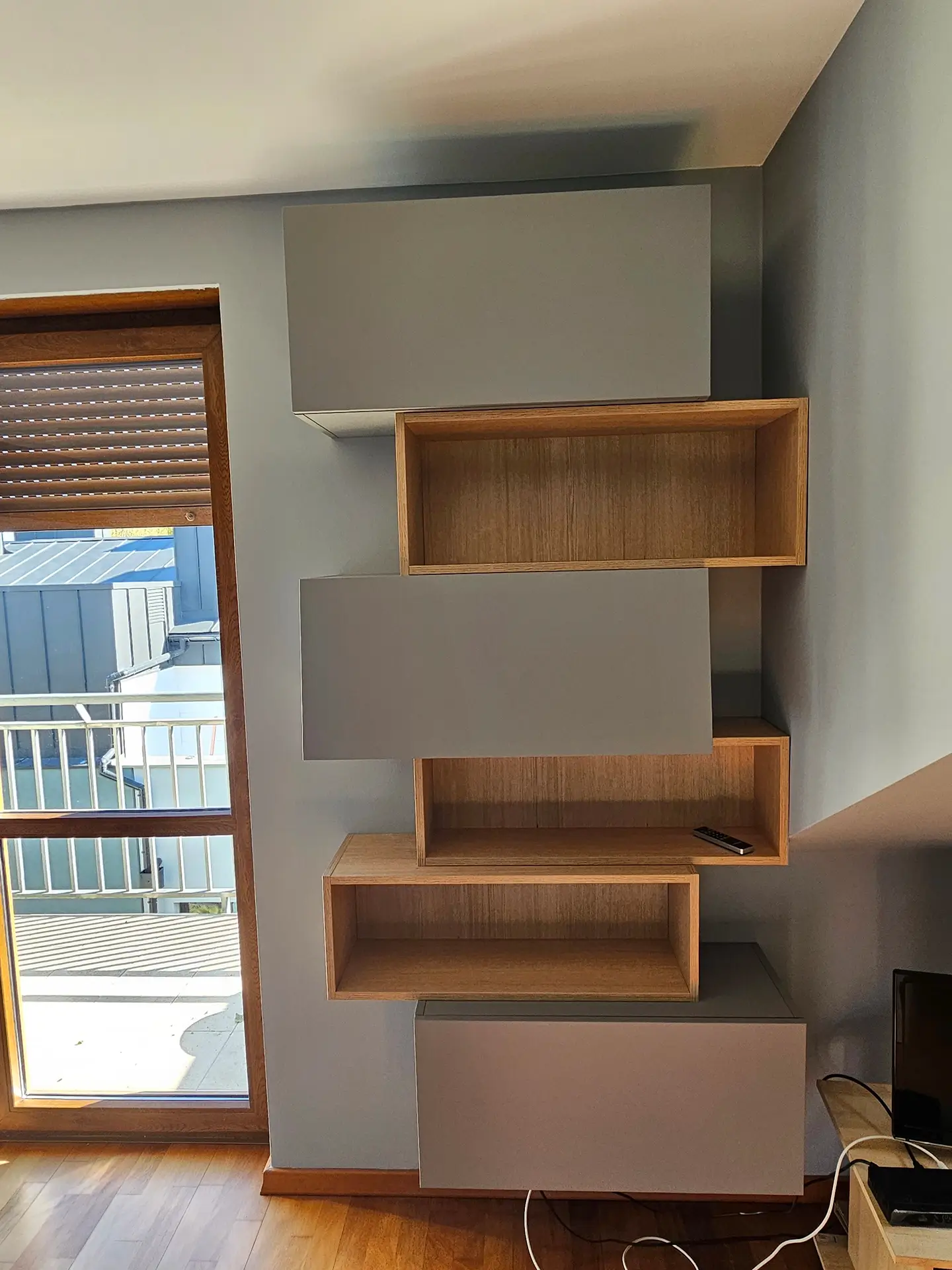 Nowoczesny, modułowy regał ścienny składający się z otwartych drewnianych półek i szarych zamykanych szafek.