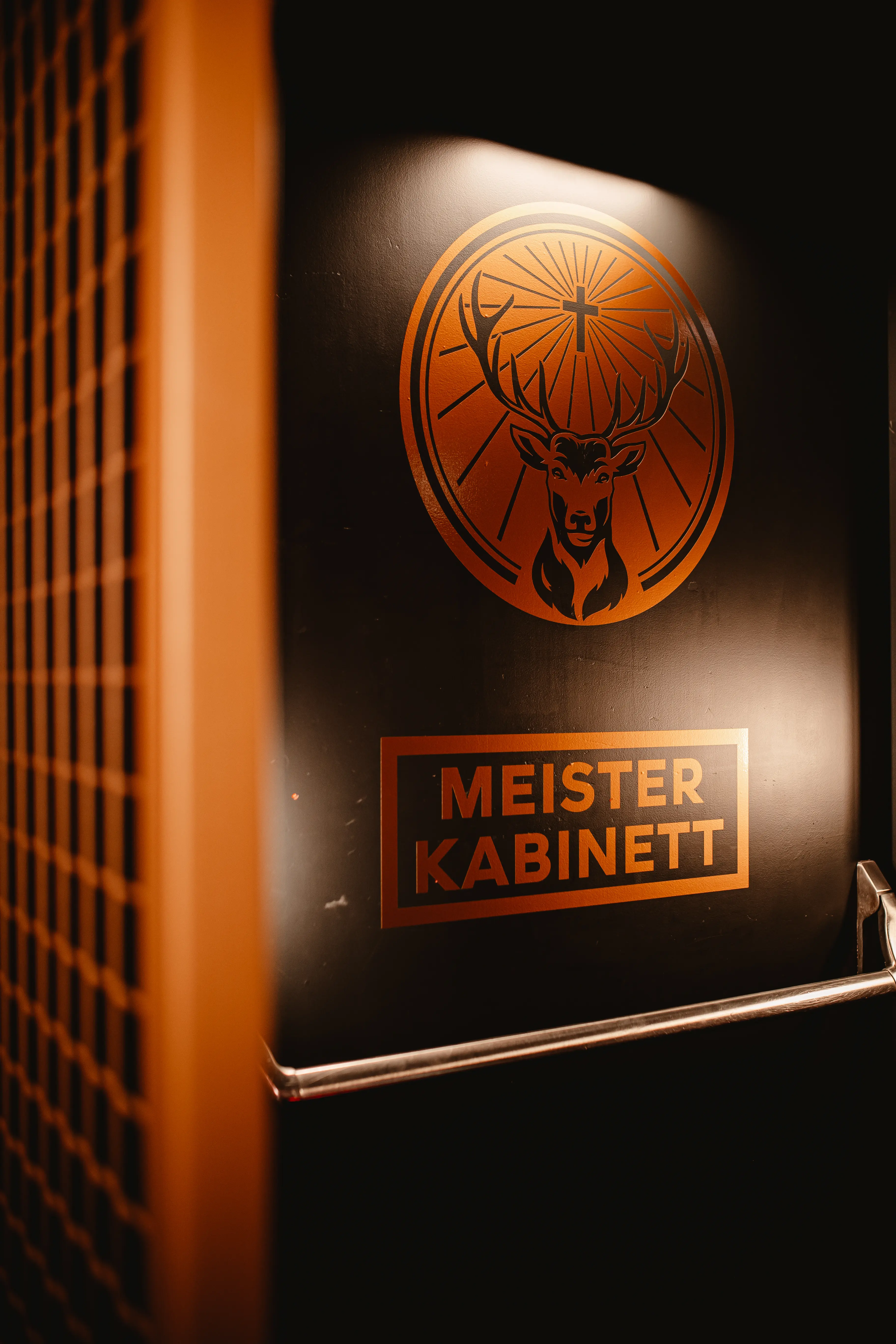 Black door with copper stag emblem and the label Meister Kabinett at JÄGERMEISTER x CALL ME DRELLA, next to an orange metal grid wall.