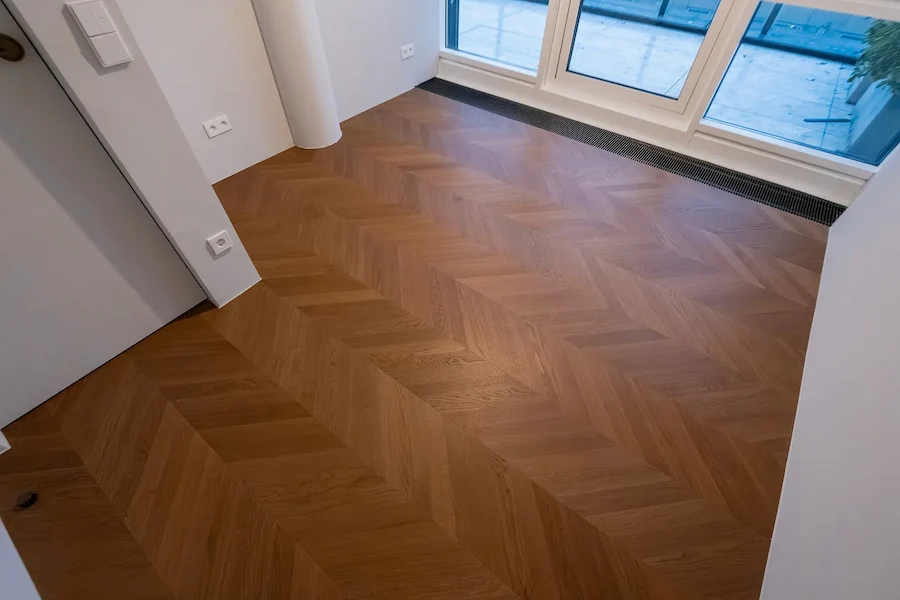 Frisch verlegter Chevron-Parkettboden in einem hellen Raum mit bodentiefen Fenstern.