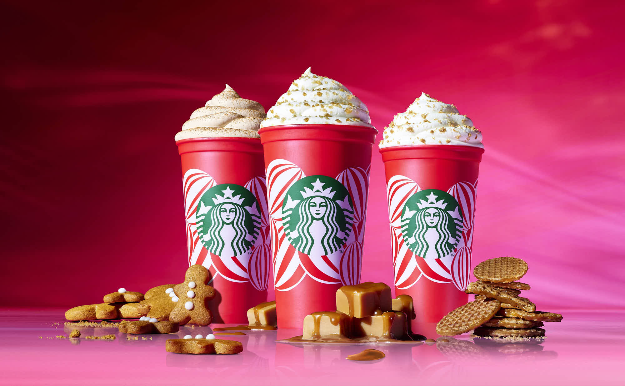 Three colorful drinks topped with whipped cream are arranged on a vibrant pink background, surrounded by snacks.