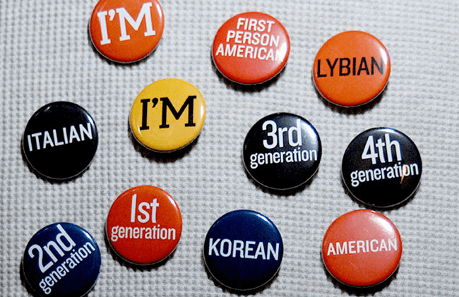 The image shows a collection of buttons on a light gray fabric background. Each button has a white text label and a distinct color. The labels include "I'm Italian", "I'm Korean", "First Generation", "Third Generation", "Fourth Generation", "Second Generation", "American", "Lybian", "Black", and "Orange". Some buttons have multiple labels. The buttons are arranged in a somewhat organized pattern, though not in a strict grid.
