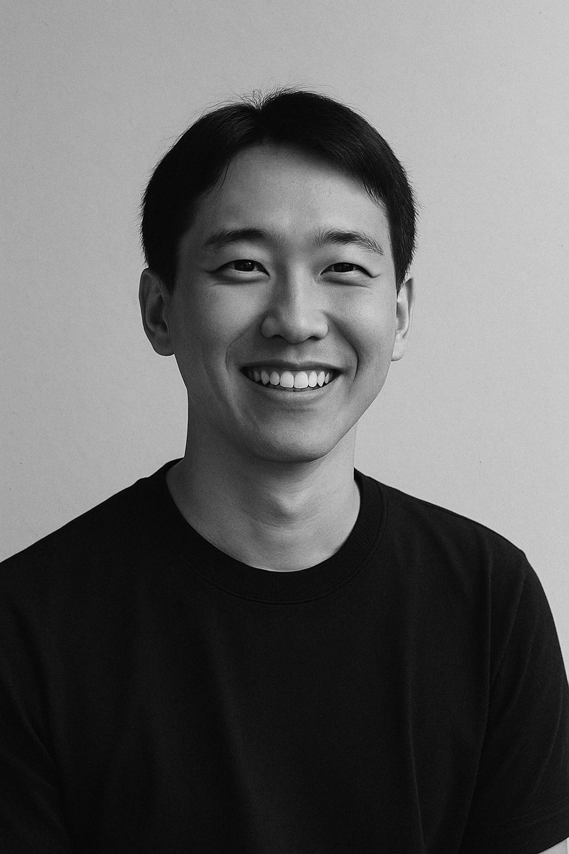 Black and white portrait of a young adult man with short, dark hair and a friendly smile, wearing a plain, dark T-shirt