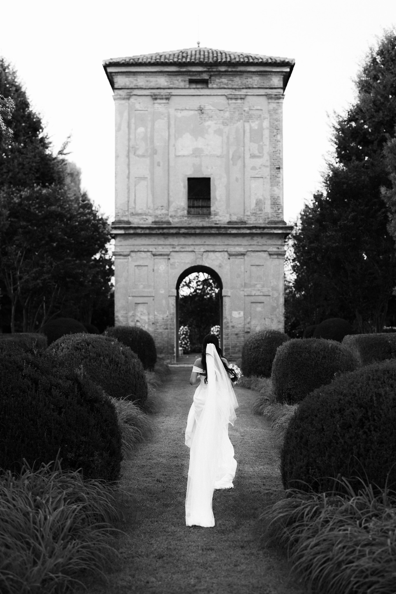Sposa cammina verso l'arco della cappella a Villa Negri, velo fluttuante, reportage matrimoniale in bianco e nero
