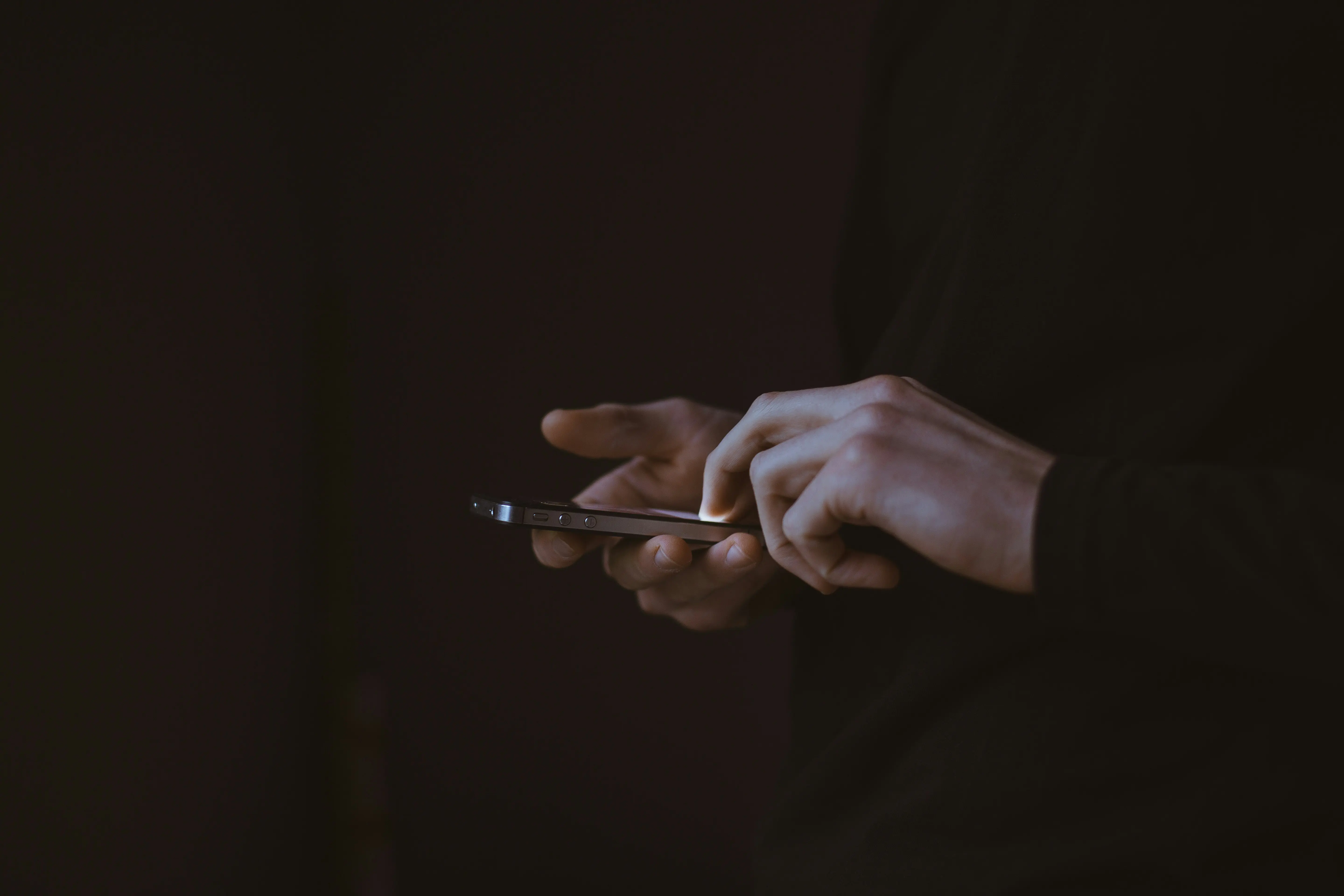 Person holding and using a smartphone with both hands against a dark background, with focus on the fingers and device.