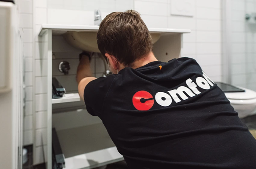 En rørlegger med en t-skjorte med merket «Comfort» på ryggen jobber under en vask på badet