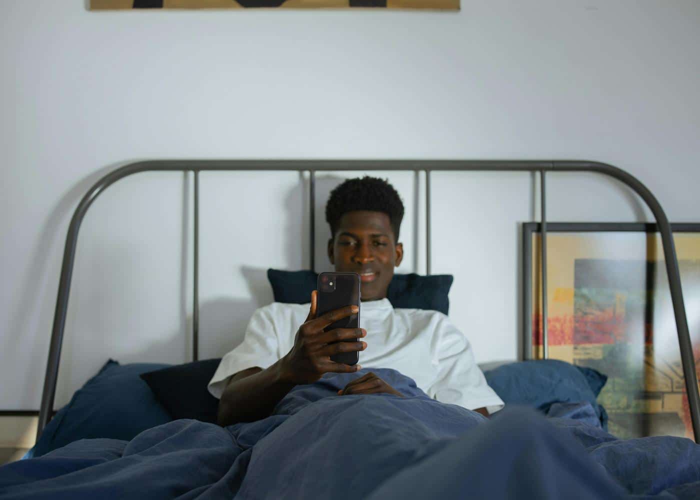 Smiling man sitting on a bed with a blue blanket on top of him looking at his phone