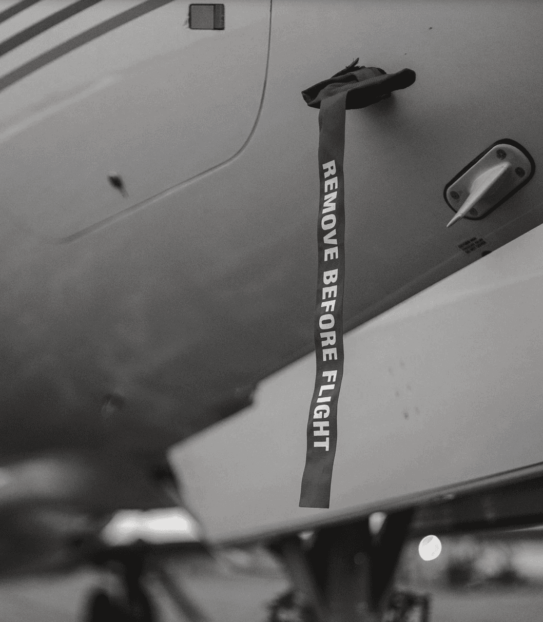 Close-up of a private aircraft underside showing a red "Remove Before Flight" safety tag attached to the landing gear