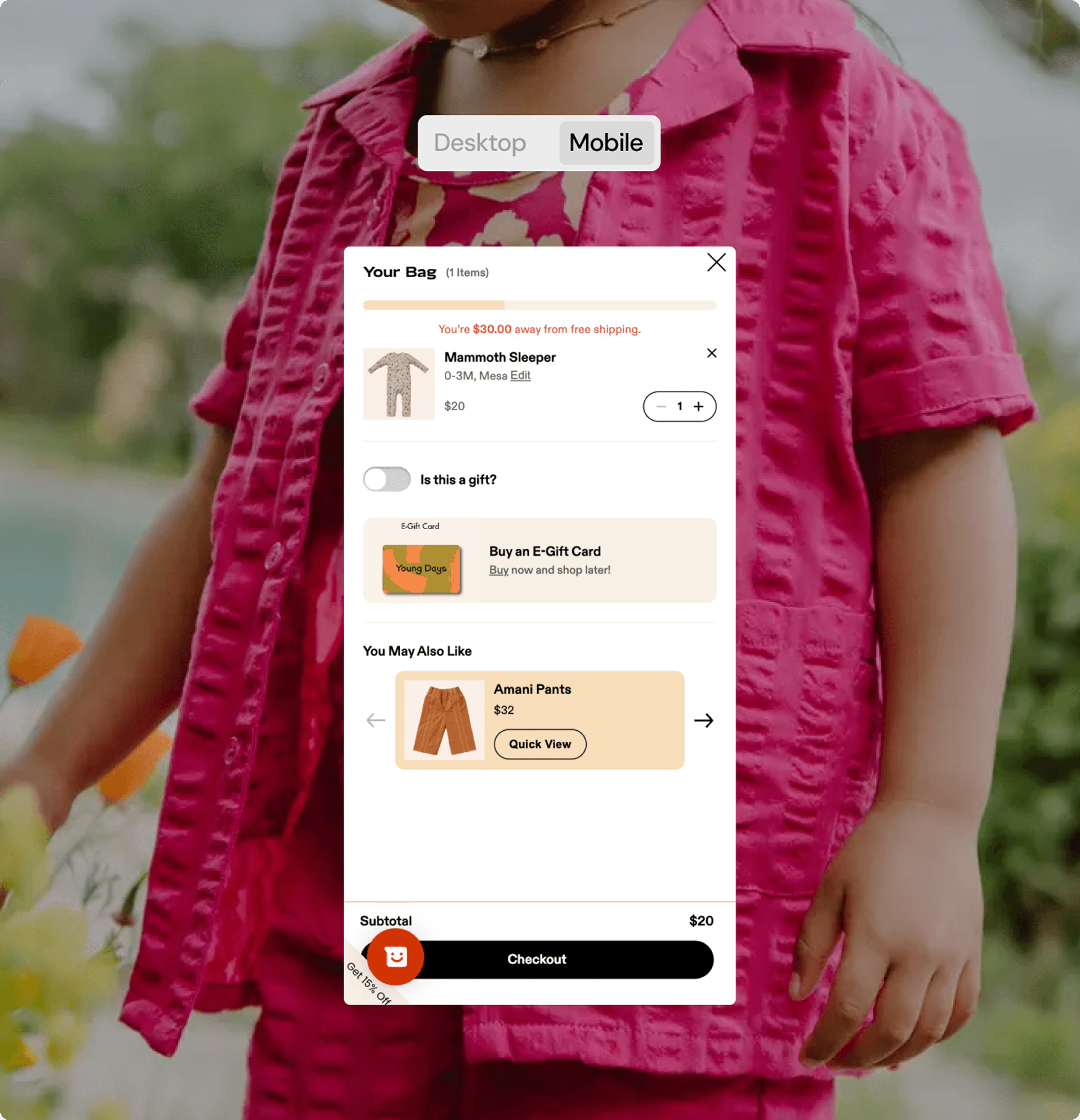 A mobile shopping cart interface is shown over a background image of a child in a bright pink outfit. The cart contains one item labeled “Mammoth Sleeper” priced at $20, with a message indicating the user is $30 away from free shipping. The interface also features gift options, an e-gift card section, and a product suggestion for “Amani Pants.” At the bottom, the subtotal is shown with a “Checkout” button. The view toggle at the top is set to "Mobile."