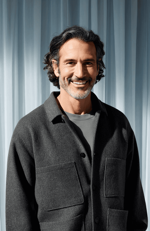 A man with long hair wearing a gray sweater, standing in a casual pose against a neutral background.
