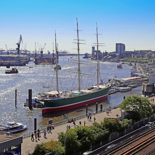 A large sailing ship docked at a busy waterfront with people walking, surrounded by smaller boats and industrial cranes in the background.