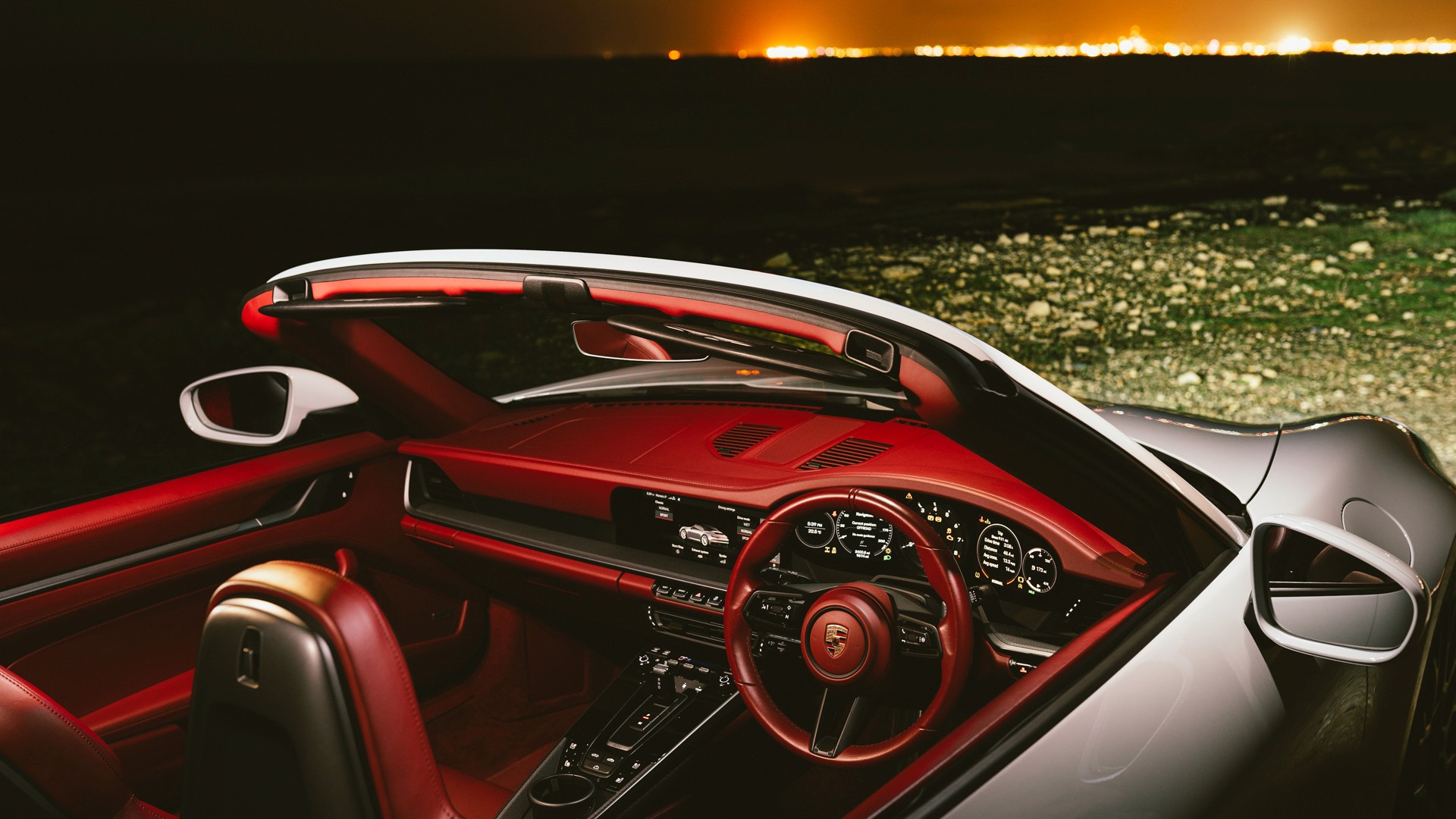 Interior of a white Porsche 911 (992) Cabriolet with the roof down, showing the red leather dashboard, door panels and steering wheel, the digital instrument cluster lit, and distant city lights glowing across dark open ground at night.