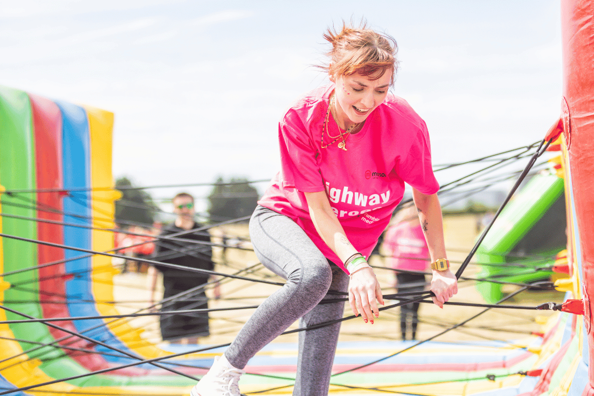 Outdoor team-building event with participants wearing matching blue shirts, taking part in inflatable obstacle courses and games, smiling and cheering, with the words 'ENTER A TEAM' in large bold white and green text across the image.