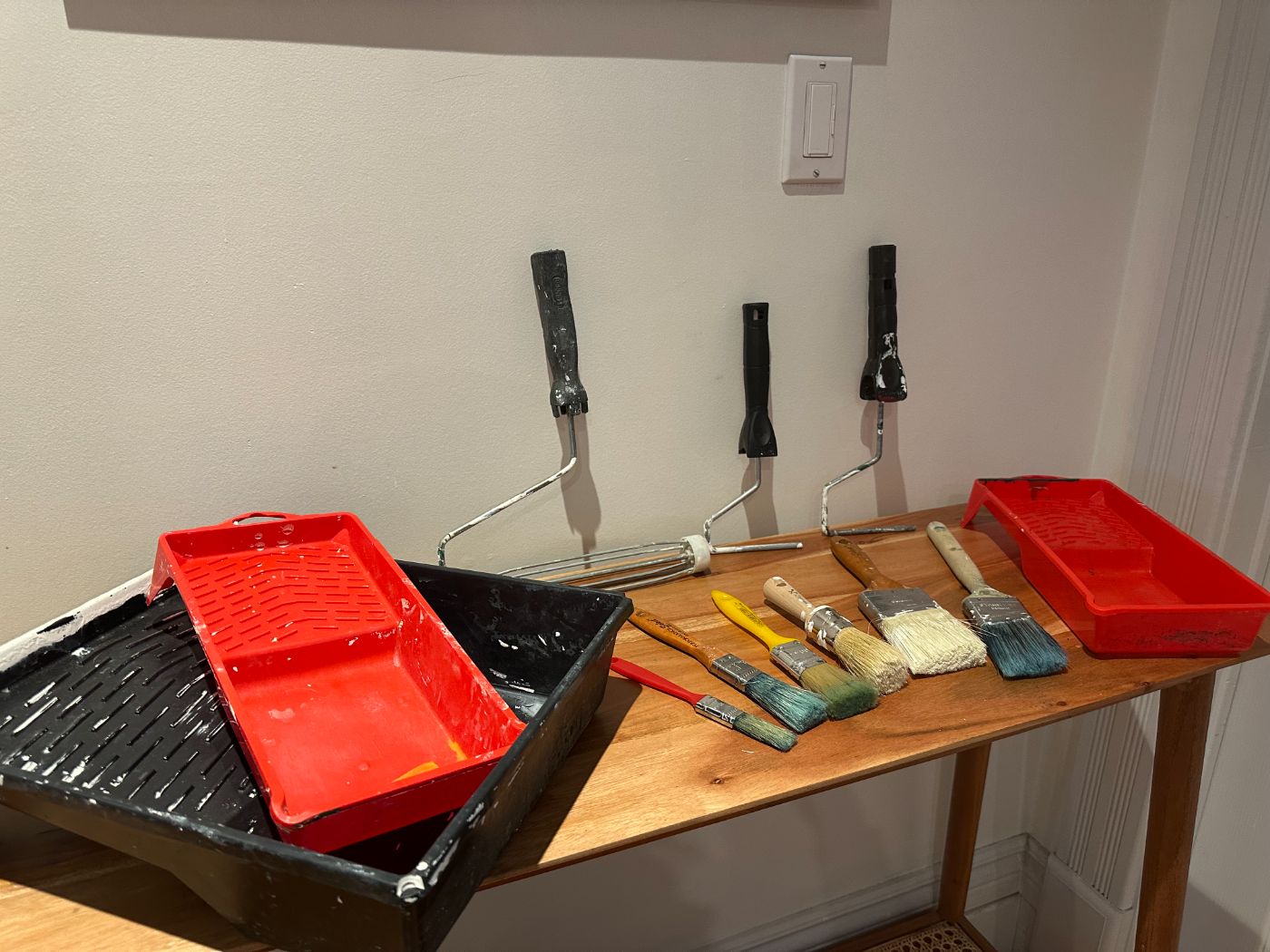Kit de peinture complet avec rouleaux, pinceaux de différentes tailles et bacs à peinture rouges et noirs, posés sur une table en bois