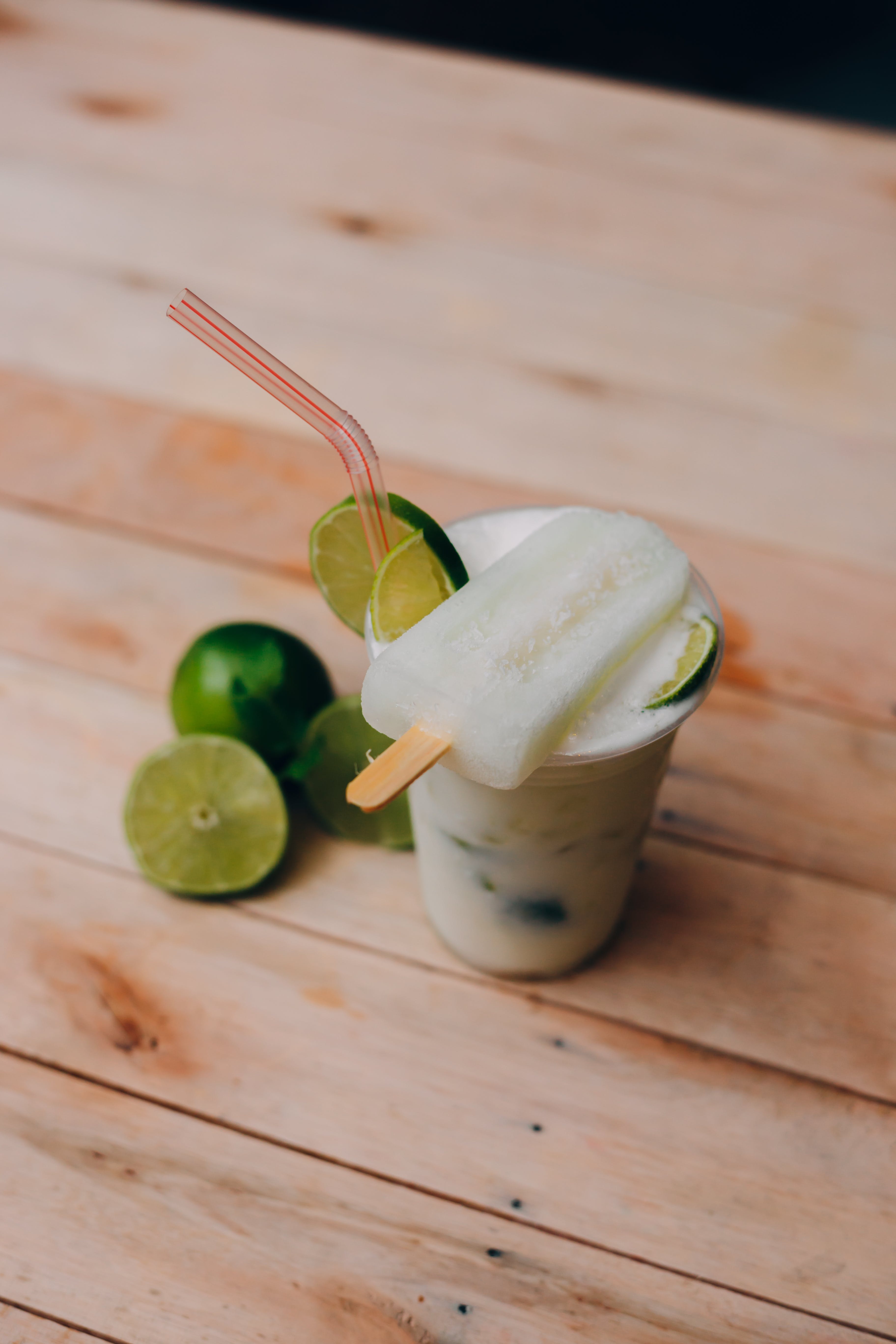 A chilled slushie drink with a popsicle laid over the lid with a straw and limes in the background