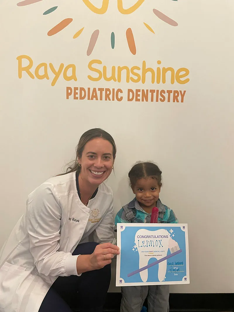 A pediatric dentist kneeling next to a smiling child, both holding a dental-themed illustration at Raya Sunshine Pediatric Dentistry