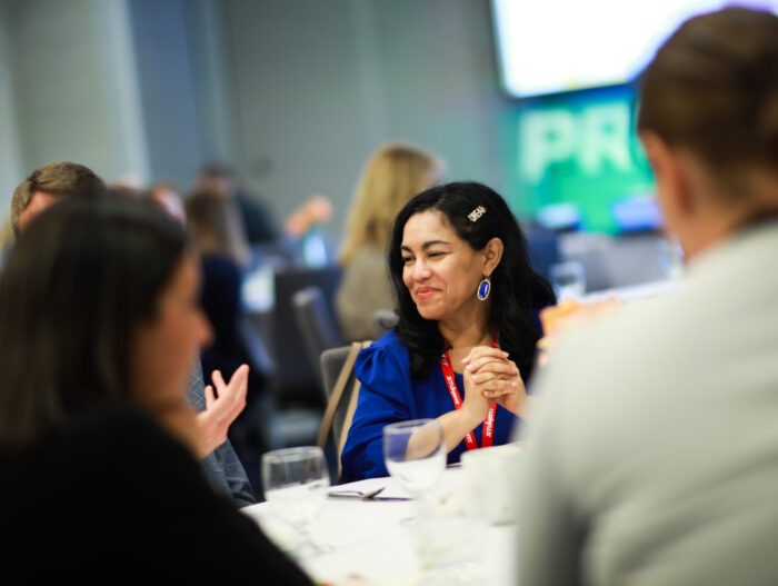Woman in blue top with red lanyard smiles while seated at a round table, engaged in conversation. Others are partially visible around her, with a blurred PROFCON sign in the background.