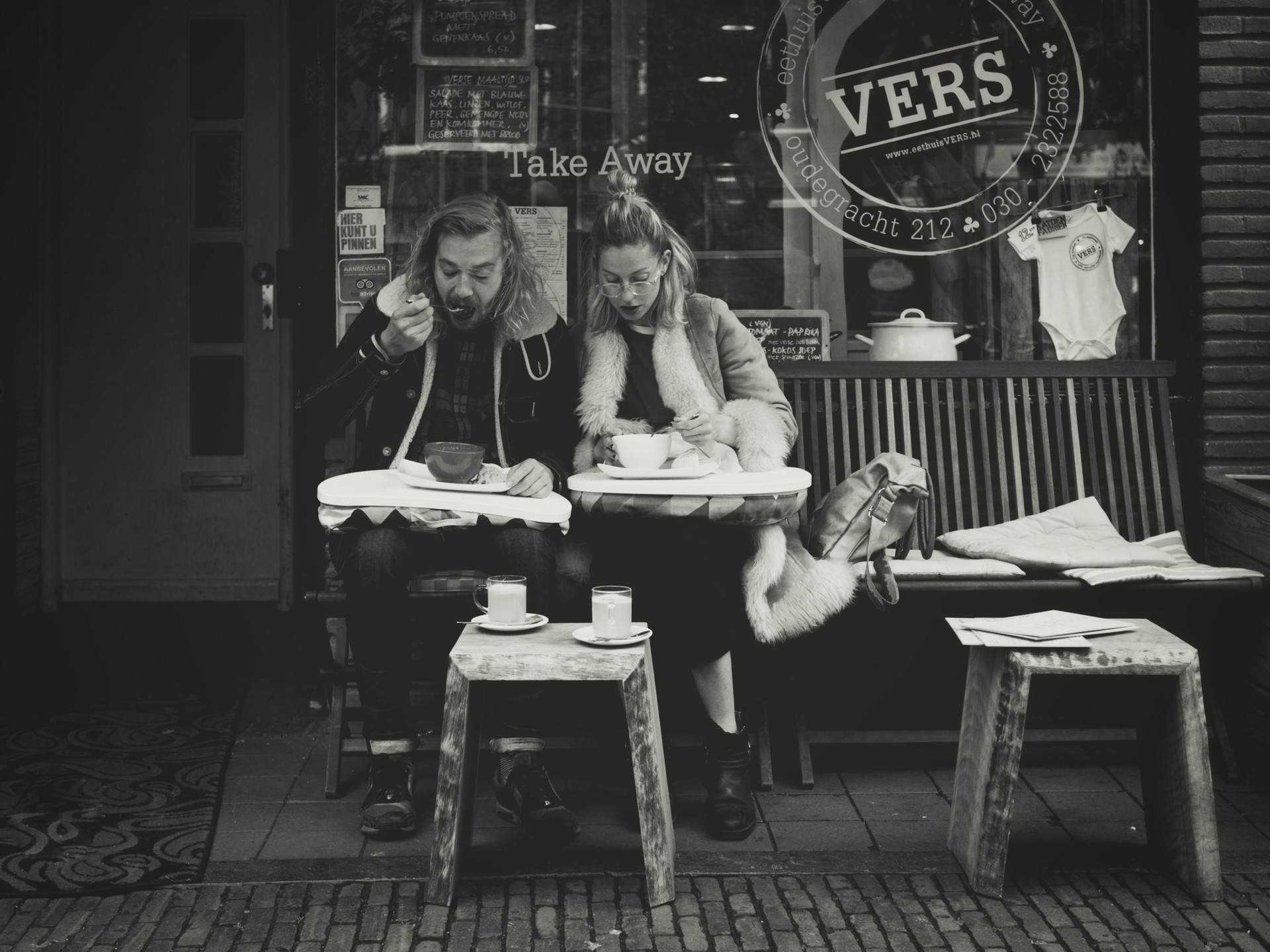 Candid black-and-white image of two people sharing a quiet meal at outdoor café tables on a city sidewalk, framed by the café’s window signage and reflective glass.