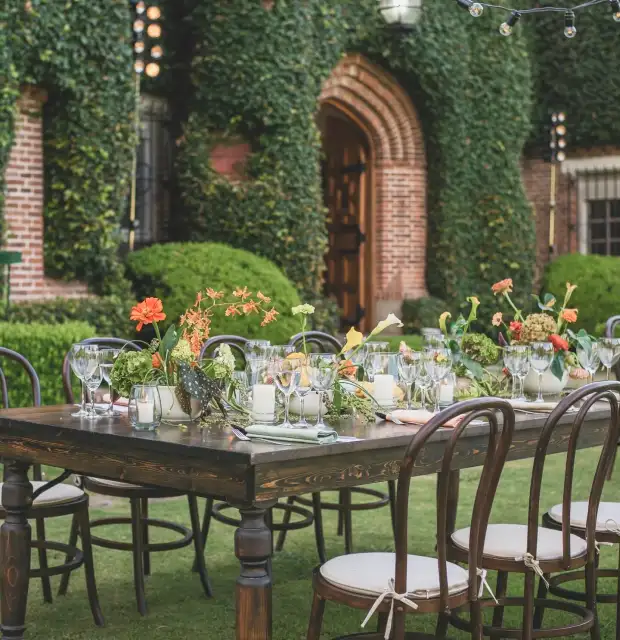 Mesa exterior decorada con flores naranjas y verdes frente a fachada con hiedra para evento elegante al aire libre, transmitiendo calidez y armonía natural.