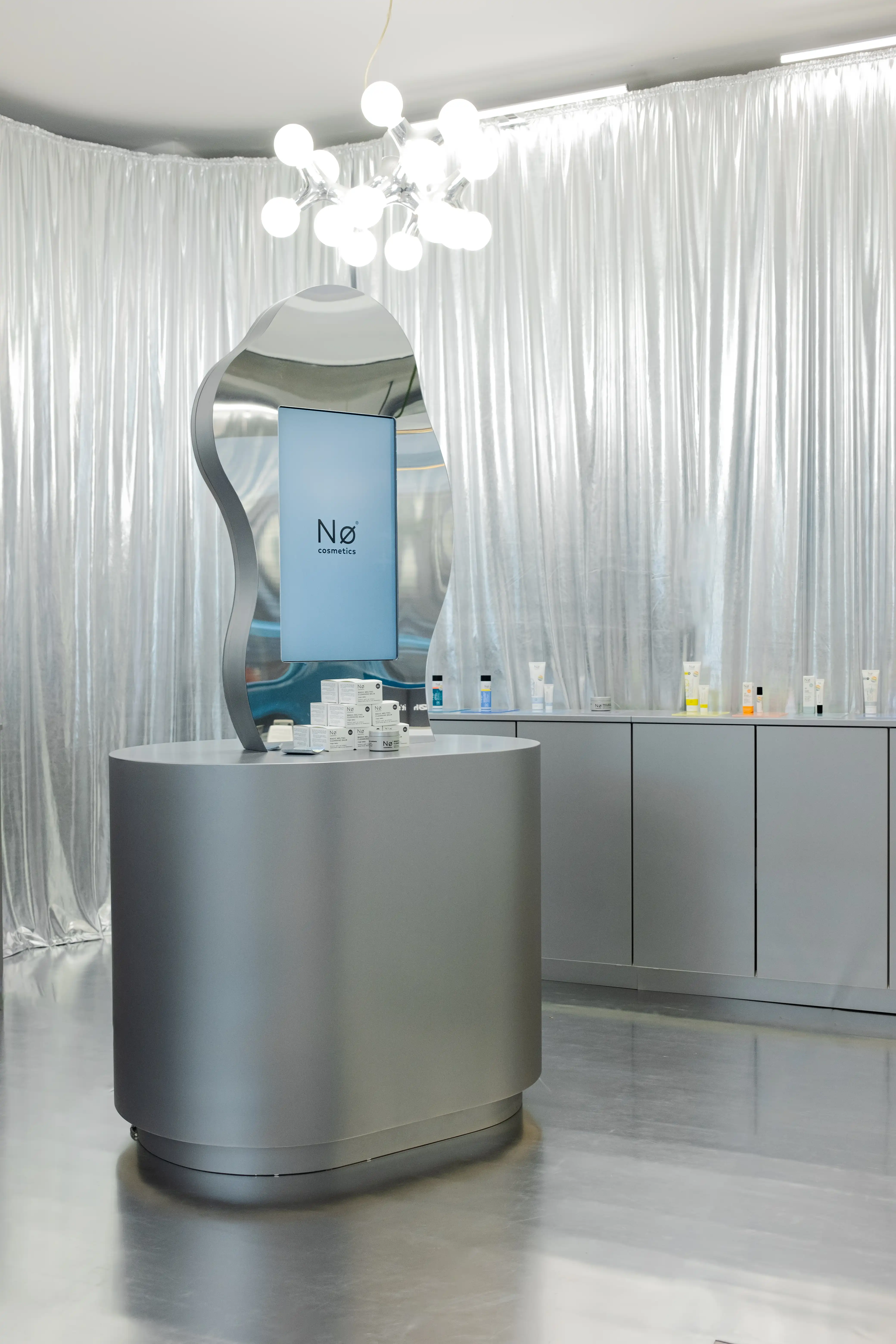 Curved chrome island with wavy screen and sphere chandelier amid silver curtains at the NØ Cosmetics pop-up.