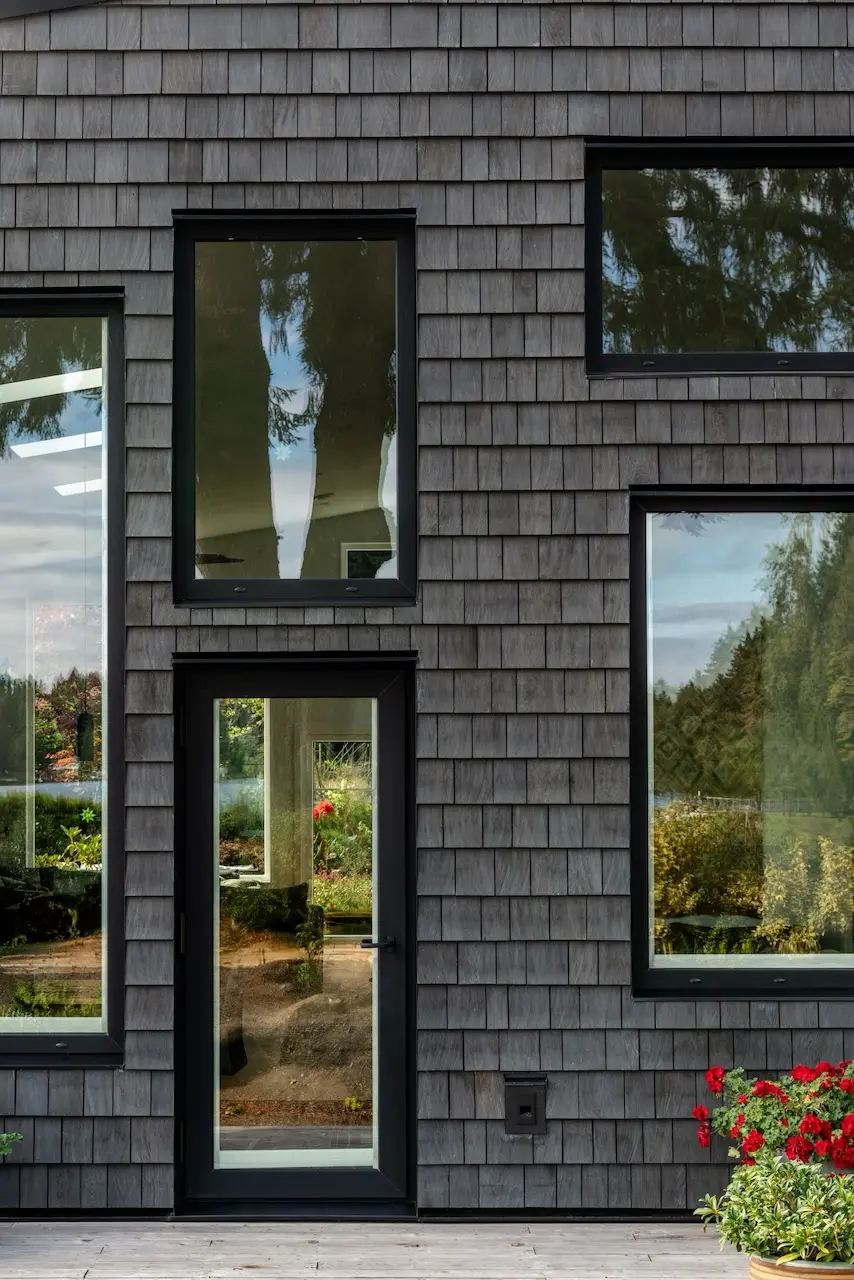 Modern two-story home with large glass windows, warmly lit interiors, and a balcony. Surrounded by trees and a manicured lawn at dusk. Cozy ambiance.