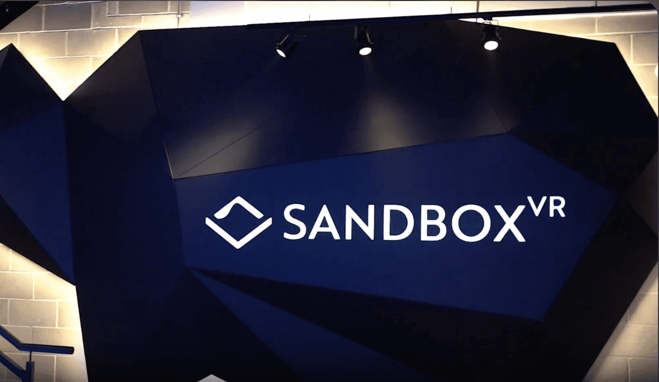 Beleuchtete Wand mit dem Logo von „SANDBOX VR“ in einem modernen, geometrischen Design. Das Logo ist weiß auf dunklem Hintergrund, und Spotlichter heben es hervor. Links führt ein Treppengeländer nach oben.