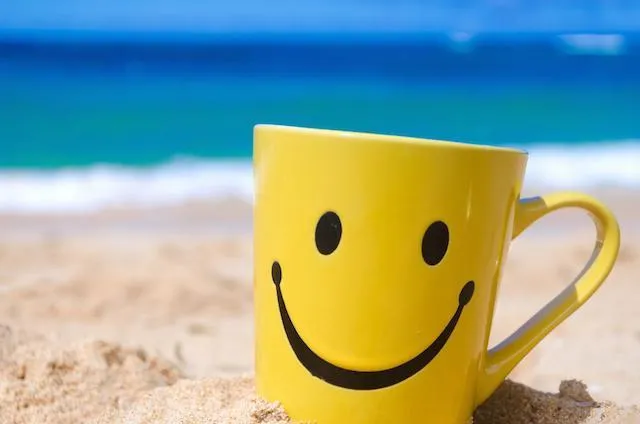 A bright yellow coffee mug with a black smiley face drawn on it sits in the sand on a beach, with the blue ocean and gentle waves in the blurred background, symbolizing relaxation and happiness.