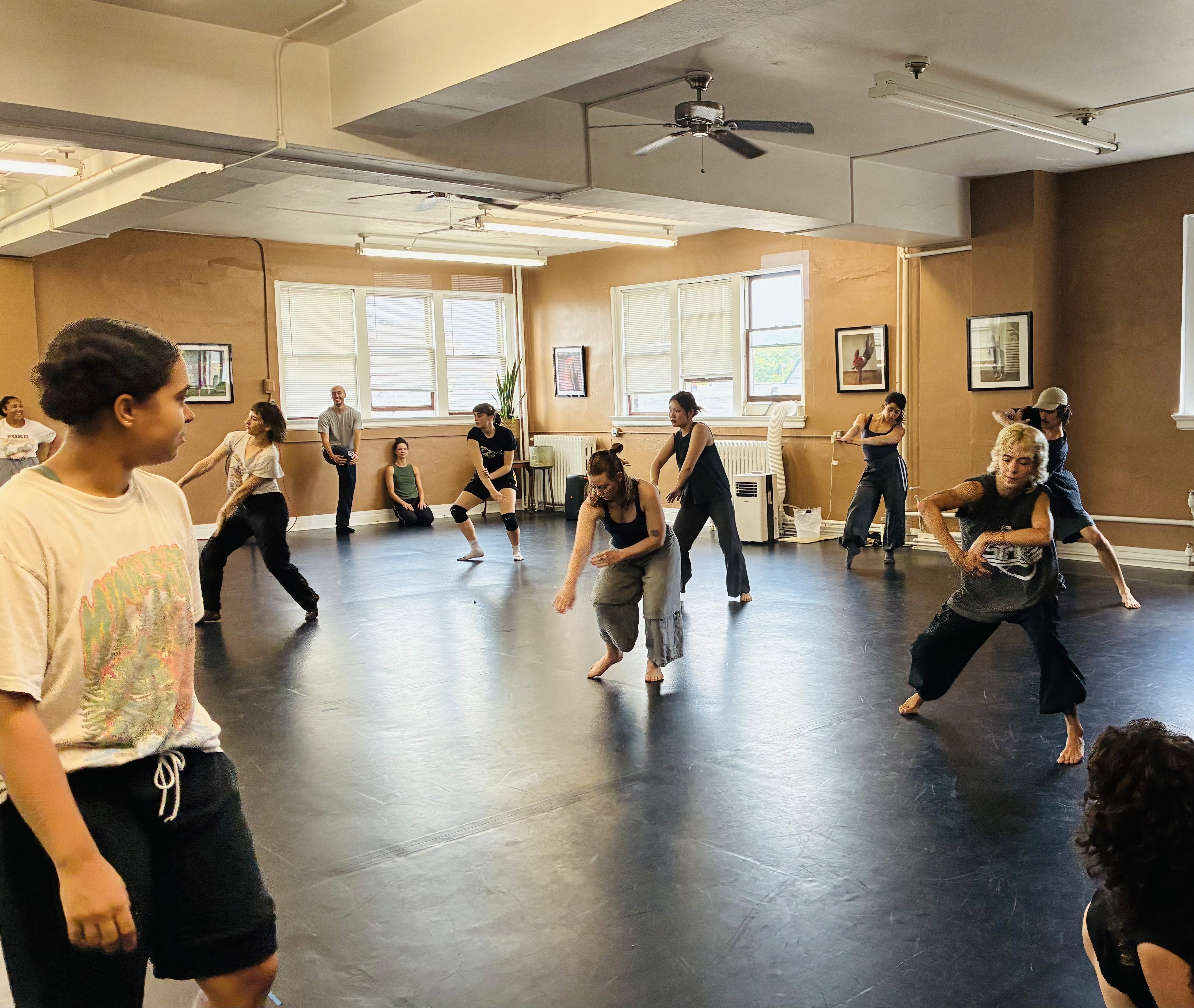 A group of contemporary dancers in a rehearsal session inside a bright dance studio.