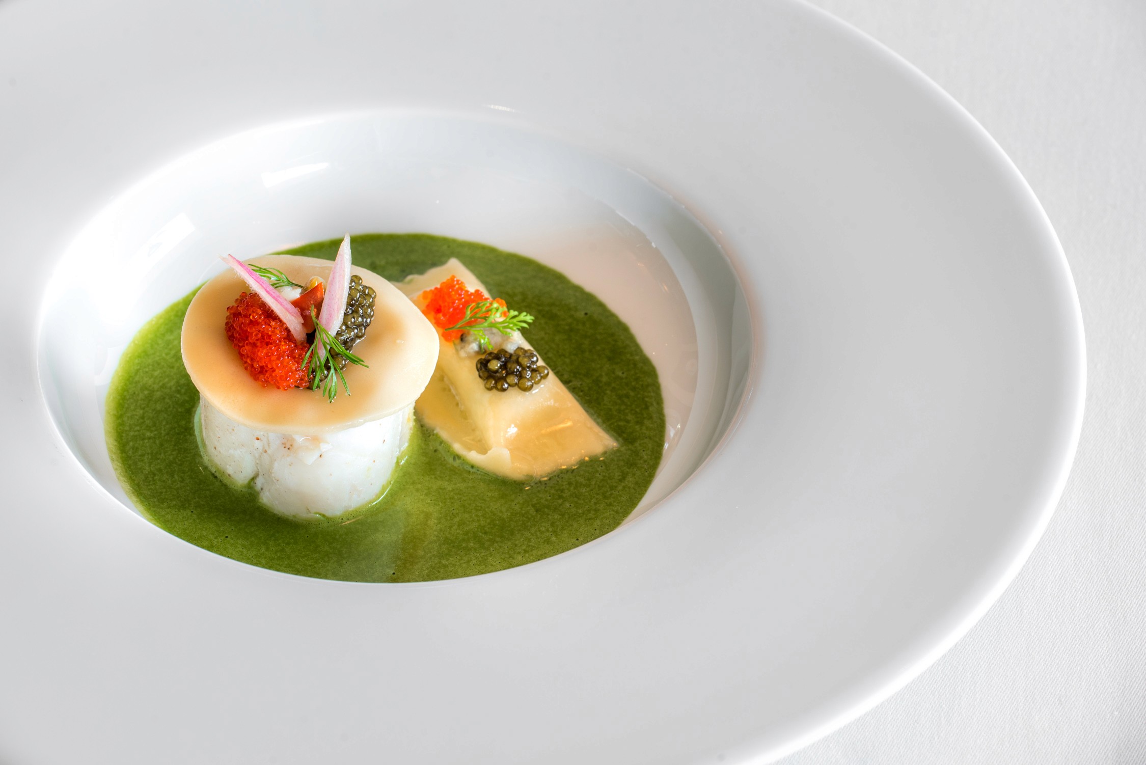 A gourmet dish featuring a seared scallop topped with a cherry tomato and herbs in a green sauce inside a white bowl.