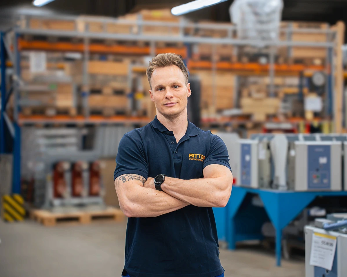 Mitarbeiter von RITTER Starkstromtechnik in der Fertigung: Er steht mit verschränkten Armen vor modernen Produktionsanlagen und Schaltschränken in einer Industriehalle.