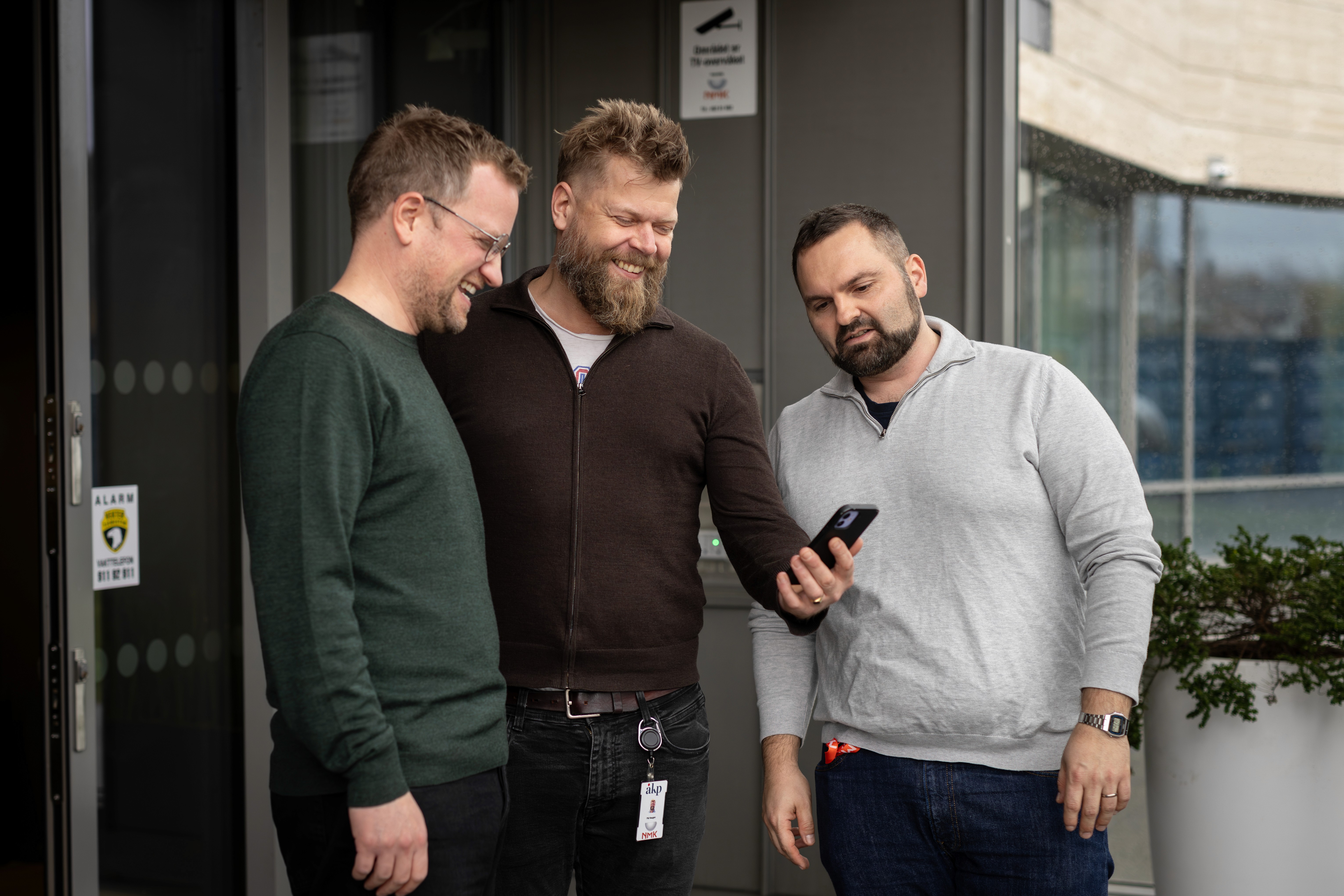 Three men stand outdoors, looking at a smartphone together, smiling and engaged in conversation.
