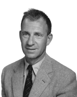 A black-and-white portrait of a man with a relaxed yet professional expression, wearing a tweed jacket, dress shirt, and tie. His slightly tousled hair and approachable demeanor convey a mix of intellect and casual confidence, characteristic of mid-20th-century style.