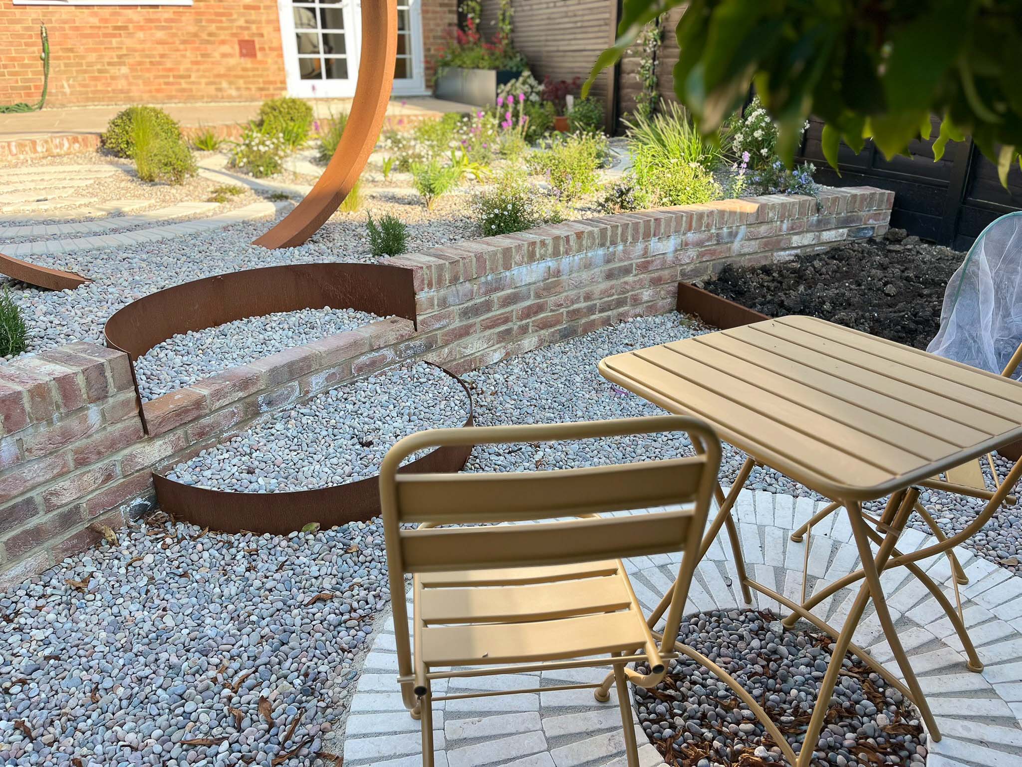 A quaint outdoor seating area with a small wooden table and chairs, surrounded by gravel and greenery.