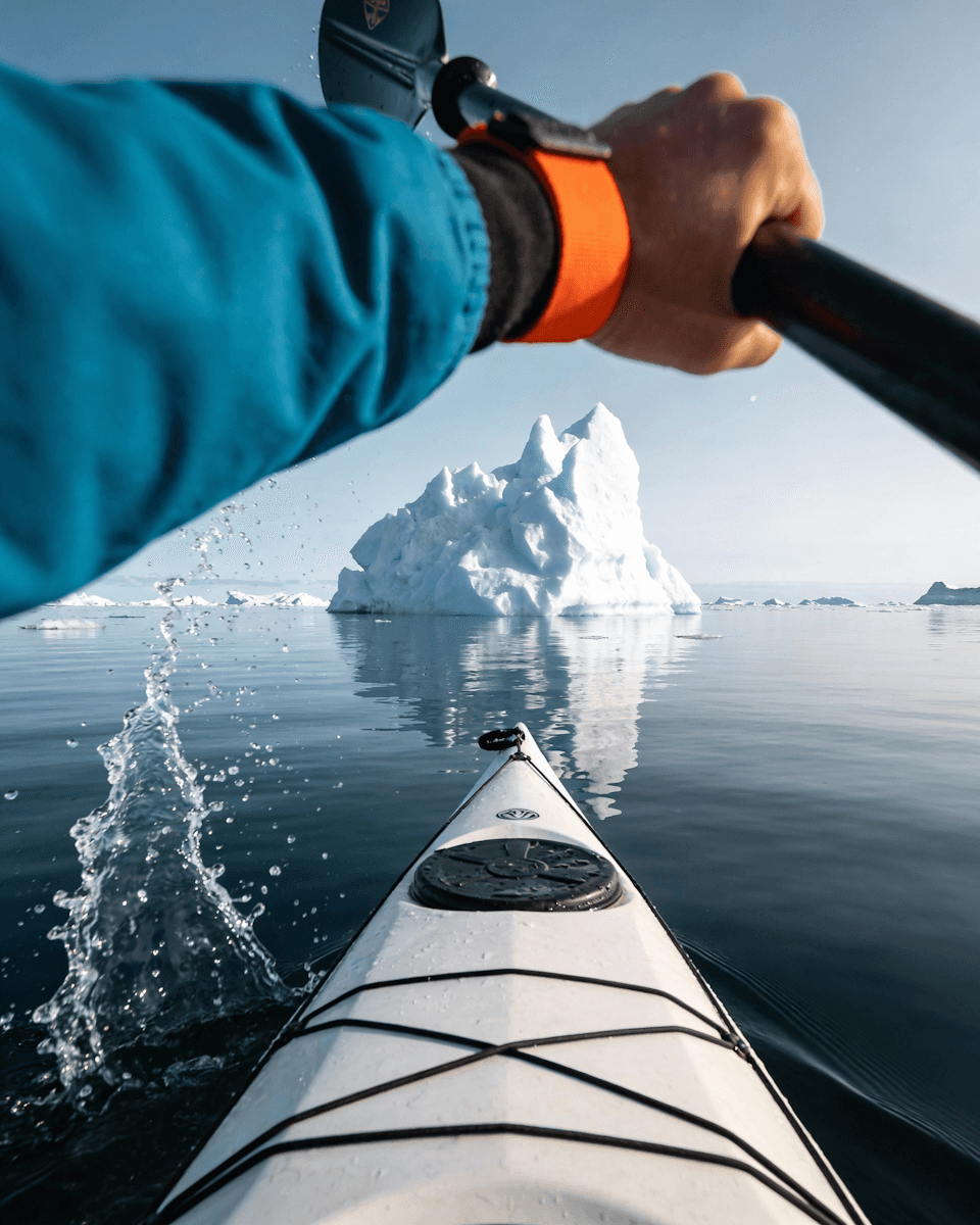 A person paddles a kayak towards an iceberg, surrounded by calm water and a clear blue sky.
