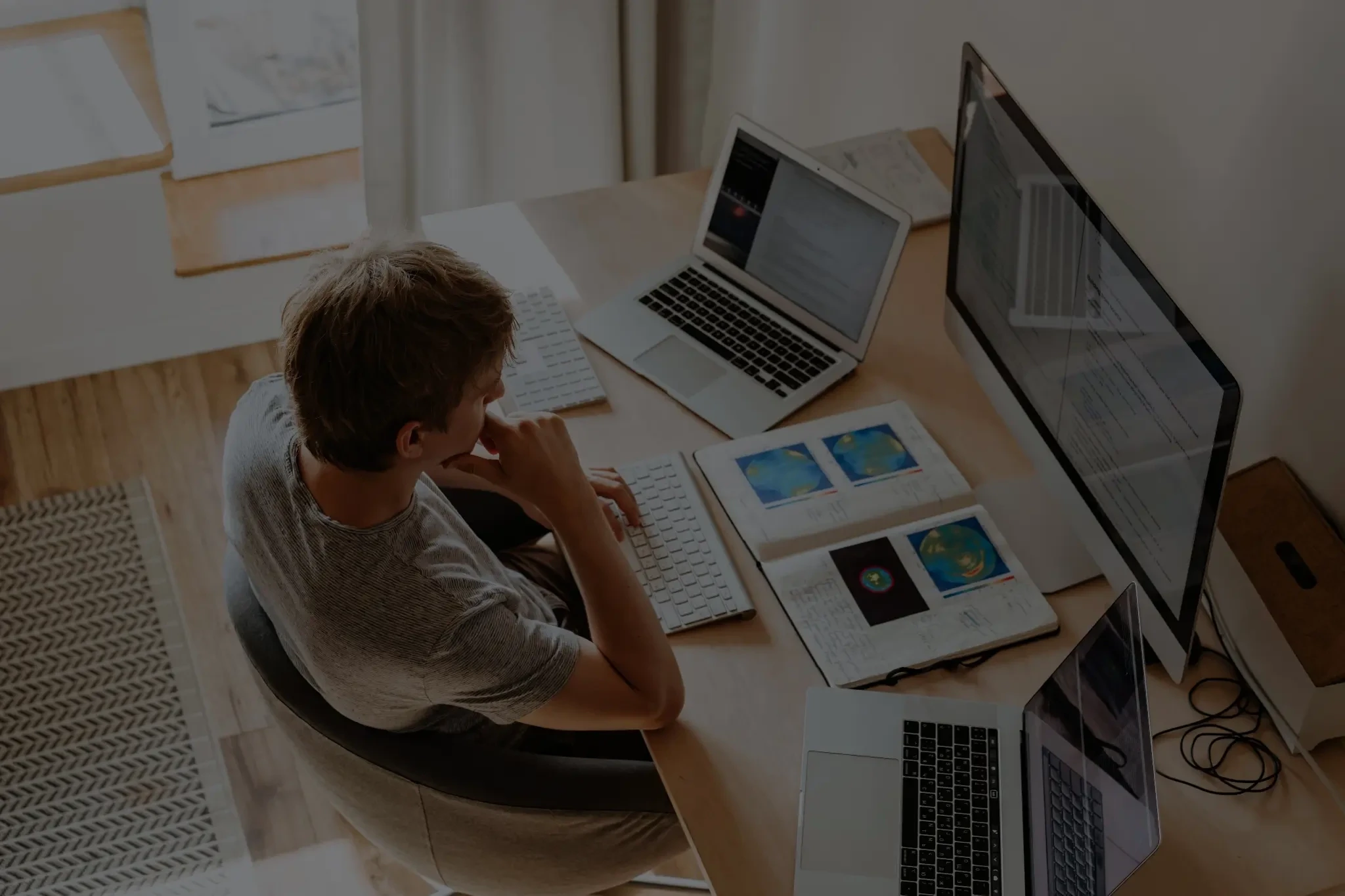 Person working at a desk with multiple laptops, a desktop monitor, documents, and data charts.