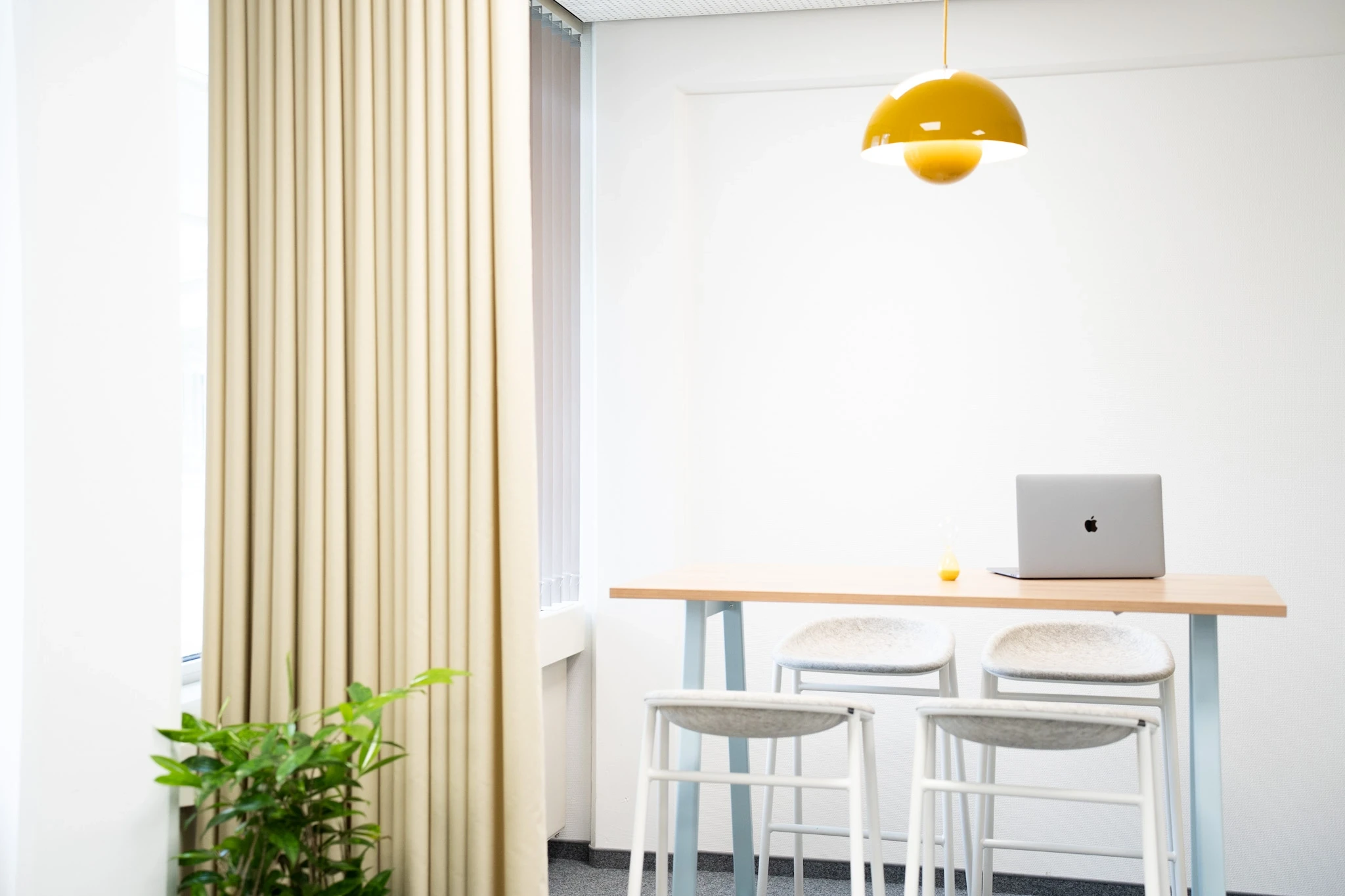 Standing desk corner at NFON with a wooden high table on light blue legs, grey felt bar stools, a mustard-yellow Flowerpot pendant lamp, beige curtain and a potted plant.