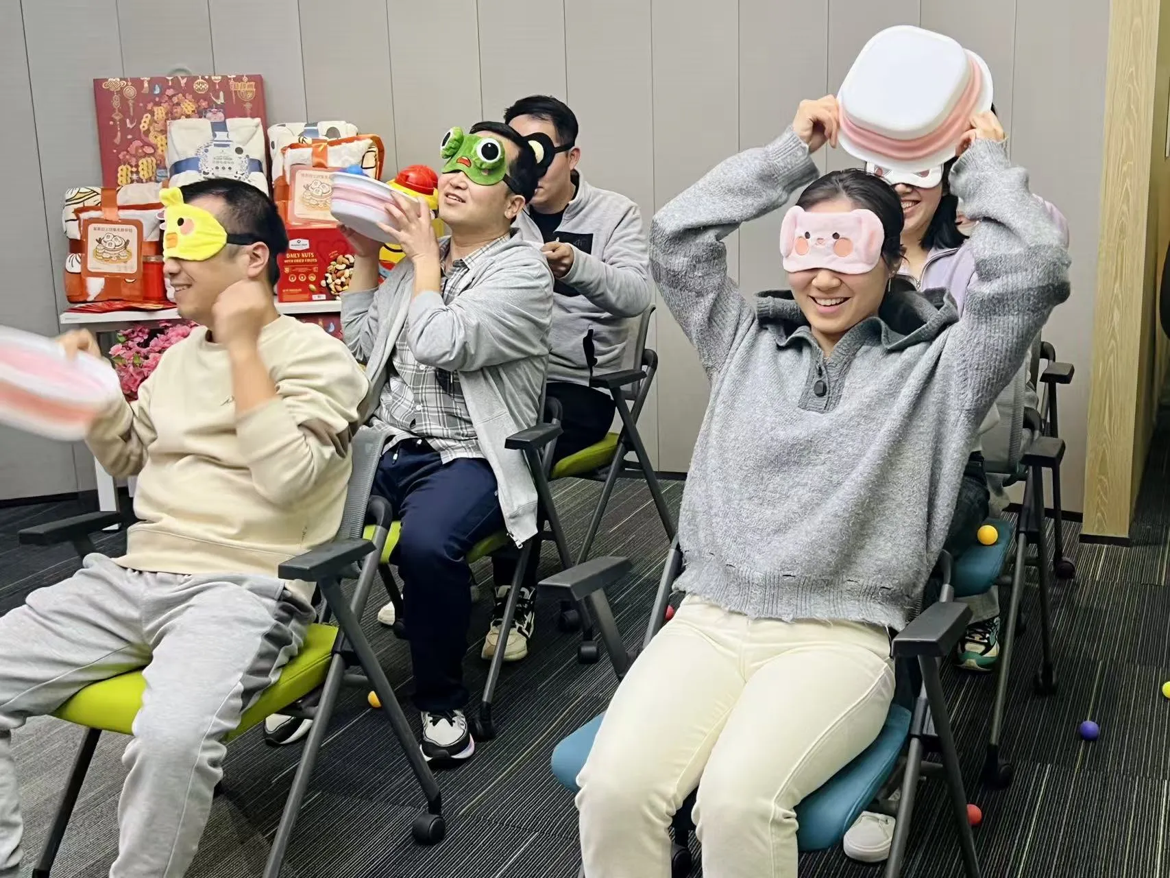 A group of people sitting in chairs, engaging playfully, some wearing hats or headgear, smiling and laughing.
