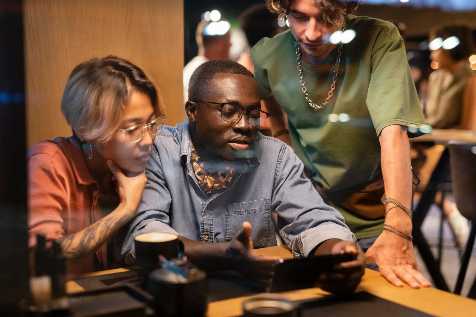 Three people gathered around a table, looking intently at a smartphone, in a cozy, dimly-lit cafe setting.
