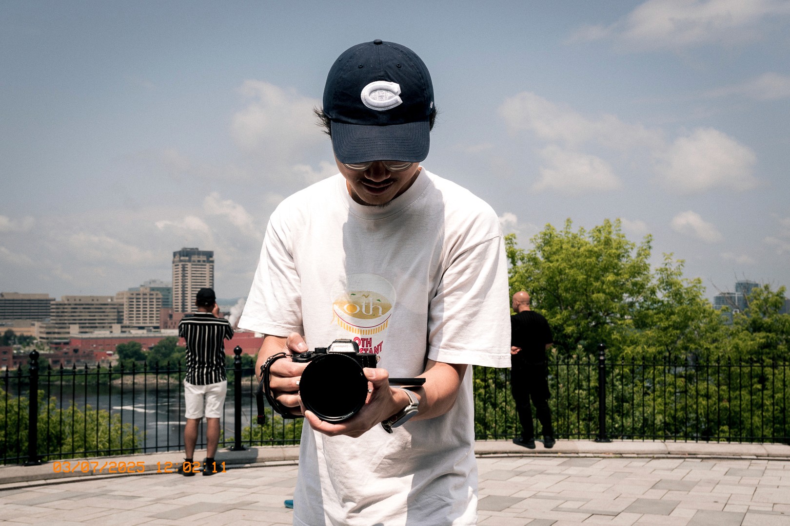 Un homme en t-shirt blanc avec logo "Oth" et casquette noire à logo "C" prend une photo. Vue urbaine avec fleuve, bâtiments et deux personnes en arrière-plan.