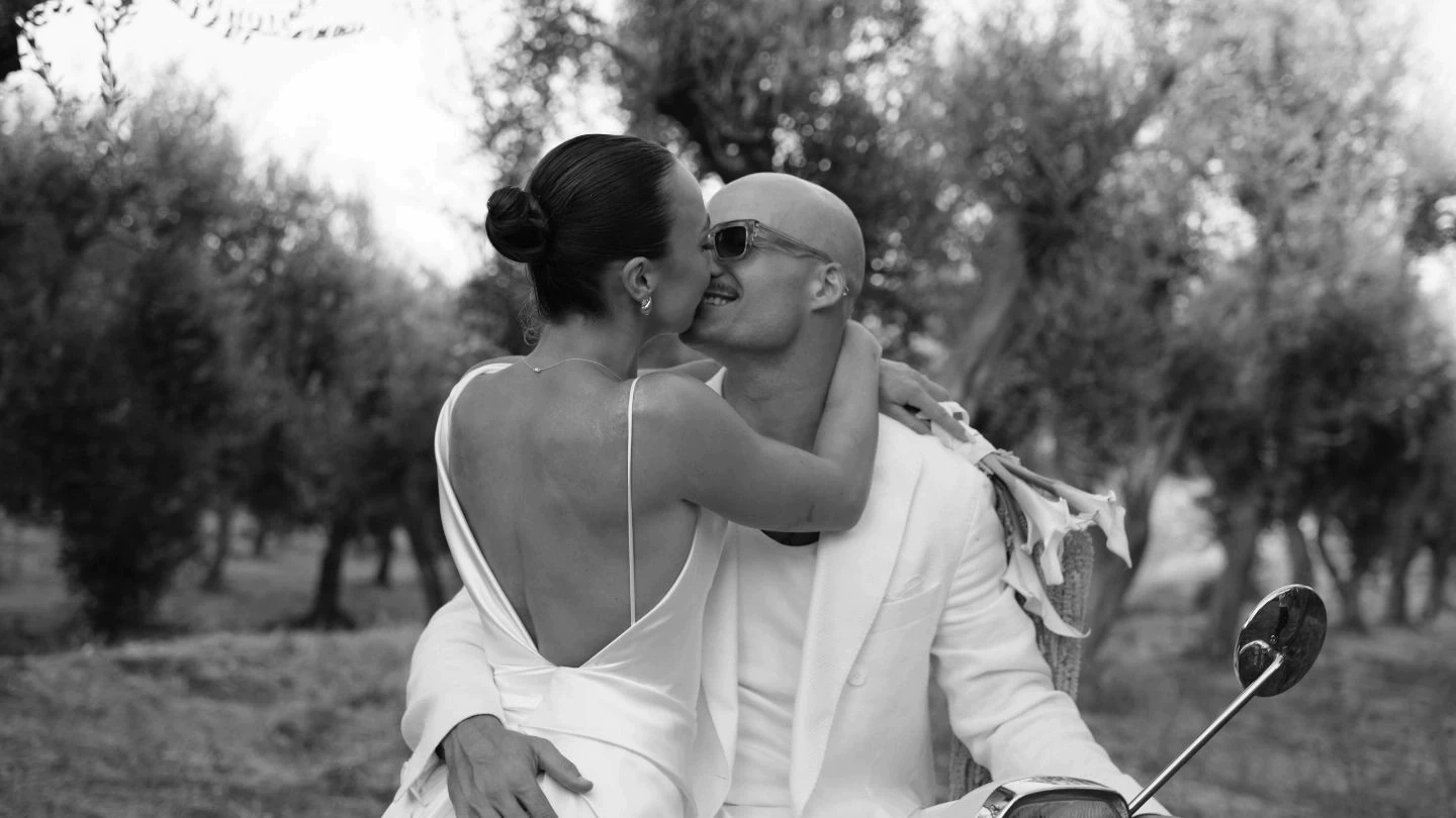 A couple sits closely in a scenic olive grove, both dressed in elegant white attire, with the man holding a golf club and the woman tenderly embracing him.