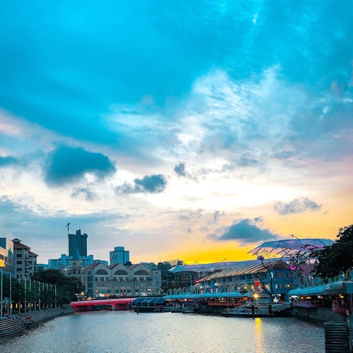 A riverside scene at sunset with buildings, bright umbrellas, and a blue sky with scattered clouds.