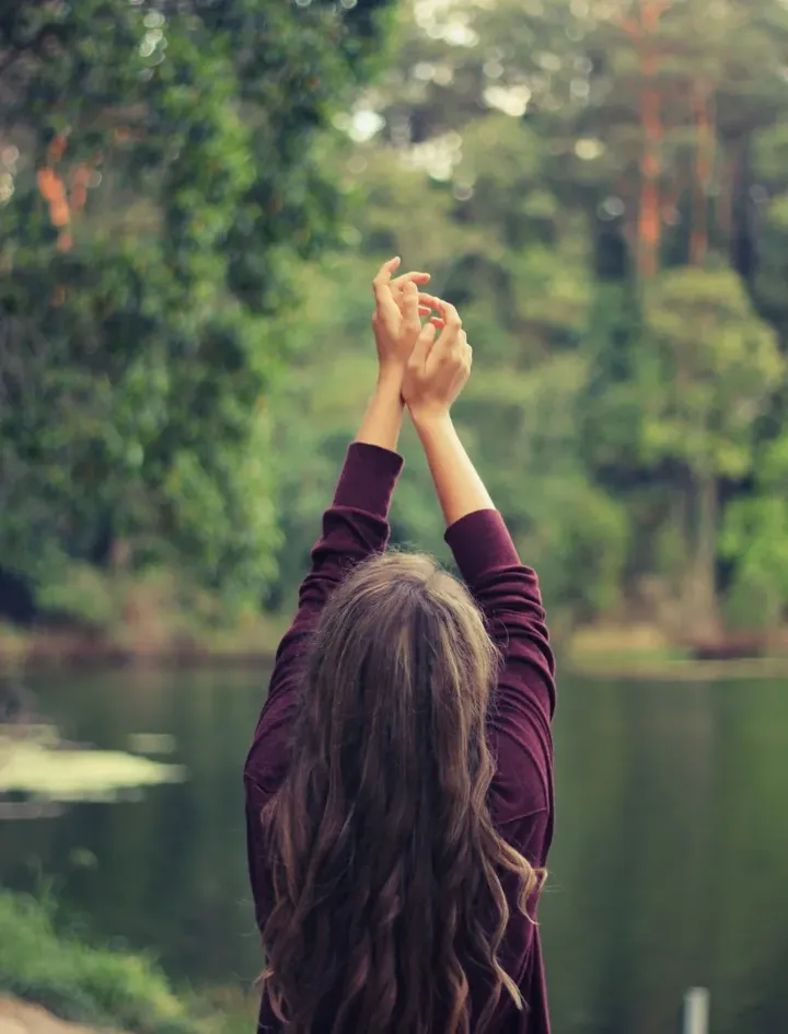 A woman stretching her arms toward the sky beside a forest lake.